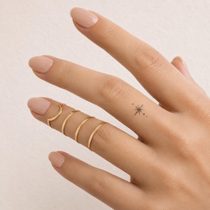 May include: A set of four thin gold rings worn on one finger, featuring a variety of smooth bands and one chevron-shaped band. A small, delicate black ink tattoo of a four-pointed star with tiny dots is visible on the adjacent finger.