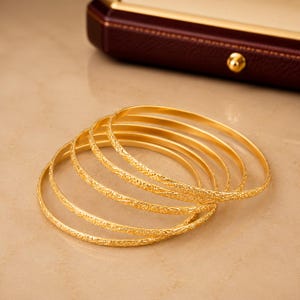 Può includere: Un set di cinque bracciali rigidi circolari e sottili in oro impilati insieme. Ogni bracciale presenta un delicato motivo inciso ripetitivo sulla superficie esterna, creando un aspetto strutturato. I bracciali hanno una finitura lucida e riflettente.