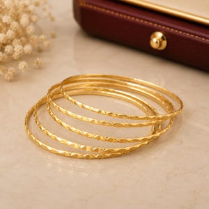 May include: A set of five thin, circular gold bangles. Each bangle features a repeating faceted texture along the outer surface, creating a series of small, reflective geometric shapes that catch the light. The bangles are stacked together.