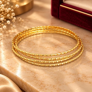 Op de afbeelding: Een set van drie dunne, ronde gouden armbanden met een herhalend textuurpatroon over het gehele oppervlak van elke band. De armbanden zijn op elkaar gestapeld en hebben een gepolijste metalen afwerking die het licht op de afgeronde randen weerkaatst.