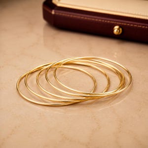 May include: A set of five thin, polished gold-tone bangle bracelets. Each bracelet features a smooth, circular band with a reflective metallic finish. The bangles are stacked together, showcasing their uniform, slender design and bright yellow gold colour.