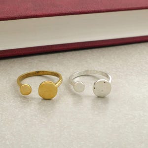 Op de afbeelding: Twee open ringen, één goudkleurig en één zilverkleurig, elk met een kleine en een grotere cirkelvormige accent. De ringen liggen op een lichtgrijze ondergrond, met een bordeauxrood boek op de achtergrond.