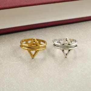 Op de afbeelding: Twee ringen met een geometrisch driehoekontwerp. De ene is goudkleurig, de andere zilverkleurig. Elke ring heeft een dubbele band en een driehoekvorm aan de boven- en onderkant. Ze liggen op een lichtgrijze ondergrond.