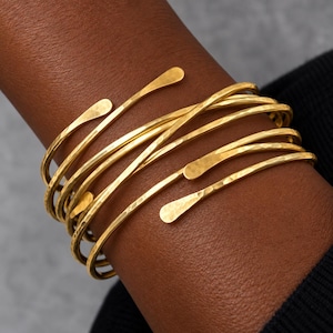 Op de afbeelding: Een goudkleurige wikkelarmband met een ontwerp van meerdere strengen en een gehamerde textuur. De armband bestaat uit verschillende dunne, flexibele metalen banden die om de pols krullen en elk eindigen in een platte, afgeronde uiteinde.