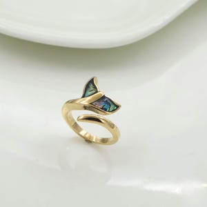 May include: A gold-toned open ring featuring a whale tail design. The top of the whale tail is inlaid with iridescent abalone shell, displaying shifting hues of green, blue, and purple. The band is smooth and tapers towards the open ends.