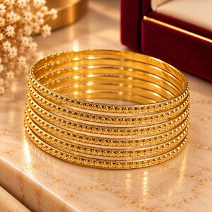May include: A set of seven gold-toned bangles stacked together. Each bangle features a polished, smooth inner band and a textured, decorative outer edge with a repeating pattern of small, raised, star-like or floral-shaped metallic details.