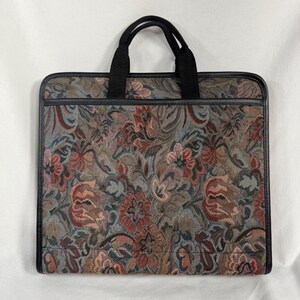 May include: A rectangular document case featuring a woven tapestry fabric with a multi-colored floral pattern in shades of muted red, blue, and green. The bag includes black fabric handles and black trim along the edges and top horizontal seam.