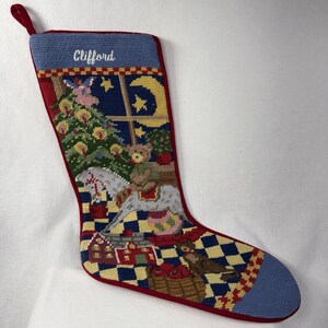 May include: A needlepoint Christmas stocking featuring a blue cuff with the name Clifford embroidered in white. The body displays a festive scene with a decorated tree, a crescent moon, a teddy bear on a rocking horse, and a dog near a basket.