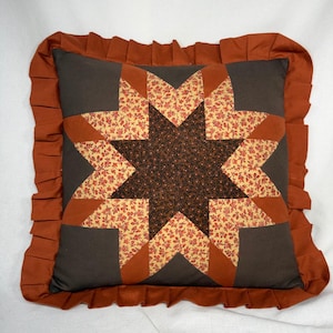May include: A square throw pillow featuring a central eight-pointed star design. The star is composed of dark brown fabric with a small floral pattern and light tan fabric with a red floral print. The pillow has a solid rust-colored ruffled border.