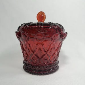 Op de afbeelding: Een dieprode glazen pot met bijpassend deksel, voorzien van een textuur met verhoogde cirkelvormige patronen en een touwachtig ontwerp rond de basis. Het deksel is bekroond met een kleine, ovale knop en het gehele object is doorschijnend.