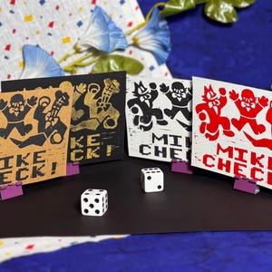 Könnte beinhalten: Vier rechteckige Karten mit einem Grafikdruck von drei Zeichentrickfiguren und dem Text MIKE CHECK! in schwarzer, goldener, weißer und roter Tinte. Die Karten stehen auf kleinen violetten Clips, davor liegen zwei weiße sechsseitige Spielwürfel.