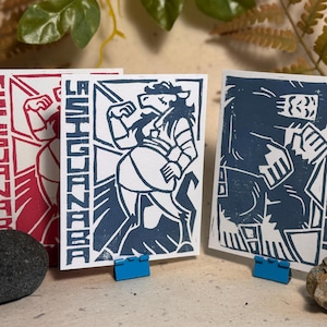 Può includere: Tre stampe artistiche rettangolari su carta bianca. La stampa a sinistra presenta una grafica rossa di La Siguanaba. Quella centrale mostra una grafica blu di La Siguanaba. Quella a destra mostra una grafica blu di Bigfoot con il testo BIGFOOT.