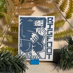 May include: A rectangular white card featuring a blue block print illustration of a stylised creature. The design depicts a large figure with textured fur and the word BIGFOOT printed vertically in bold, blocky capital letters on the right side.