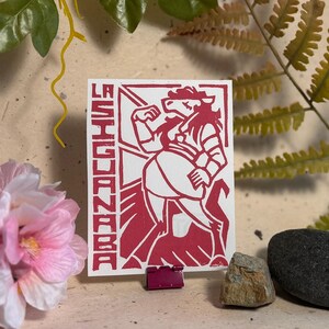 May include: A rectangular white card featuring a bold red ink print of a stylised figure with a horse-like head and human-like body. The text LA SIGUANABA is printed vertically in a blocky font along the left side of the card, which is held by a small clip.