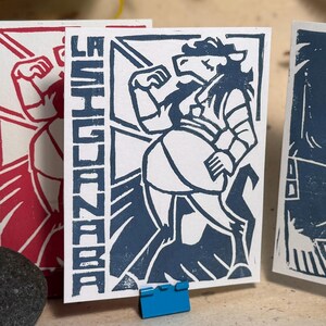 May include: A rectangular white card featuring a dark blue linocut print of a mythical creature with a horse-like head and human-like body. The text LA SIGUANABA is printed vertically in a bold, blocky font along the left side of the card.