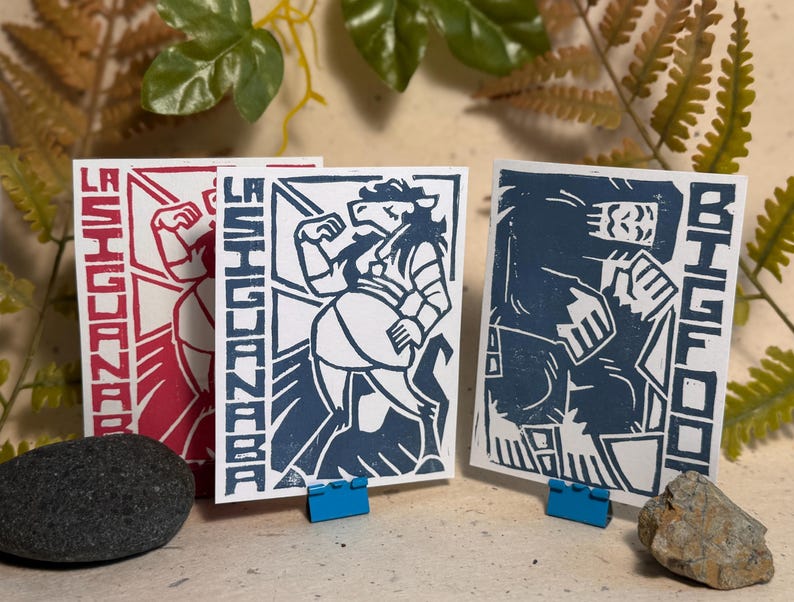 May include: Three rectangular art prints on white paper. The left print features a red graphic of La Siguanaba. The centre print shows a blue graphic of La Siguanaba. The right print displays a blue graphic of Bigfoot with the text BIGFOOT.