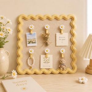 Puede incluir: Un tablero de corcho cuadrado de color beige con un marco amarillo ondulado. Seis clips blancos en forma de margarita están sujetos al tablero, sosteniendo una fotografía, un pequeño ramo de flores secas, una nota con el texto focus on the good, una pulsera de perlas, una tarjeta floral y un pequeño oso de peluche tejido a ganchillo.