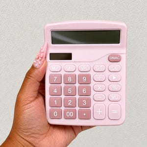 Pu&ograve; includere: Una calcolatrice da tavolo rosa chiaro con uno schermo rettangolare e una griglia di pulsanti quadrati. I tasti numerici sono rosa chiaro, mentre i tasti funzione sono di una tonalit&agrave; di rosa leggermente pi&ugrave; scura, disposti in layout.
