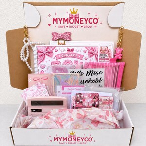 Pu&ograve; includere: Una scatola di cartone con il marchio MYMONEYCO contenente un planner di budget rosa, una calcolatrice rosa, un portachiavi di perle, un portachiavi a forma di orsetto rosa, una clip a fiocco glitterata e vari articoli di cancelleria.