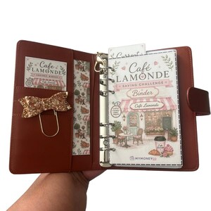May include: A brown leather-effect six-ring binder featuring a Cafe Lamonde savings challenge insert. The interior includes a gold glitter bow paperclip, a patterned side pocket, and a clear divider with a cafe illustration and text.