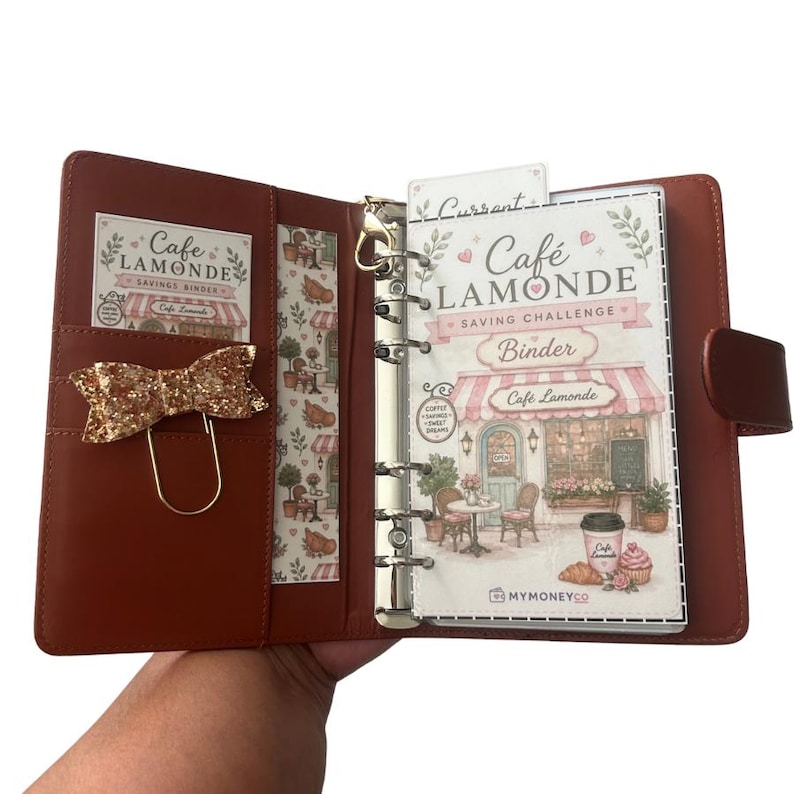 May include: A brown leather-effect six-ring binder featuring a Cafe Lamonde savings challenge insert. The interior includes a gold glitter bow paperclip, a patterned side pocket, and a clear divider with a cafe illustration and text.