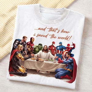 May include: A white t-shirt featuring a graphic print of various superheroes and a central figure seated at a table with the text "...and That's how i saved the world!" printed in brown script above the scene, showing a dinner setting.