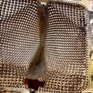 Op de afbeelding: Een vintage metalen muntportemonnee met een goudkleurig patroon van in elkaar grijpende schakels. De portemonnee heeft een scharnierend rechthoekig frame met een metallic afwerking en een fijn kettinghengsel aan de bovenkant.