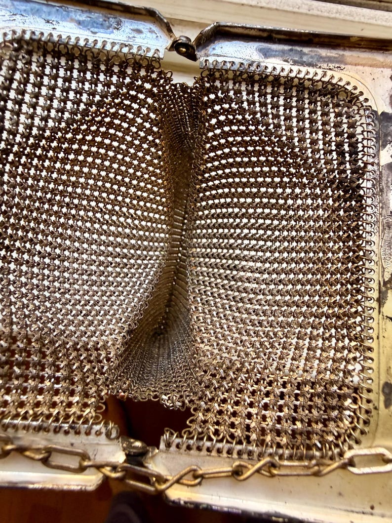 Op de afbeelding: Een vintage metalen muntportemonnee met een goudkleurig patroon van in elkaar grijpende schakels. De portemonnee heeft een scharnierend rechthoekig frame met een metallic afwerking en een fijn kettinghengsel aan de bovenkant.
