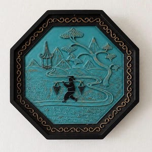 May include: An octagonal wall plaque featuring a black frame with gold scrollwork accents. The central teal panel displays a relief scene of a figure carrying baskets on a pole, a pagoda, mountains, and a tree, all rendered in black and teal.