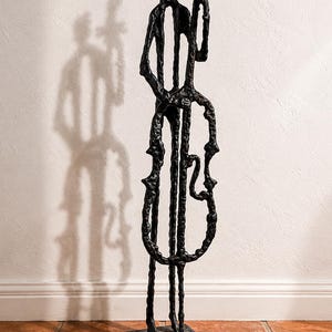 May include: A black metal sculpture of a musician playing a cello. The abstract figure stands on a square base. The sculpture's form is defined by thick, textured lines, and it casts a shadow on the wall behind it.