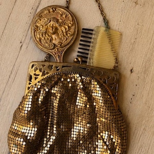 May include: A gold-toned metal mesh handbag featuring an ornate filigree frame and a chain handle. Inside the bag, a round hand mirror with a relief portrait of a woman and a small comb with black bristles are tucked into the main compartment.
