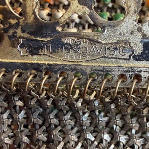 Op de afbeelding: Een vintage metalen mesh tas van Whiting & Davis Co. met een decoratief filigraan frame. Het metalen gaas bestaat uit kleine, in elkaar grijpende zilverkleurige schakels met subtiele oranje en groene emaille accenten zichtbaar in het weefsel.