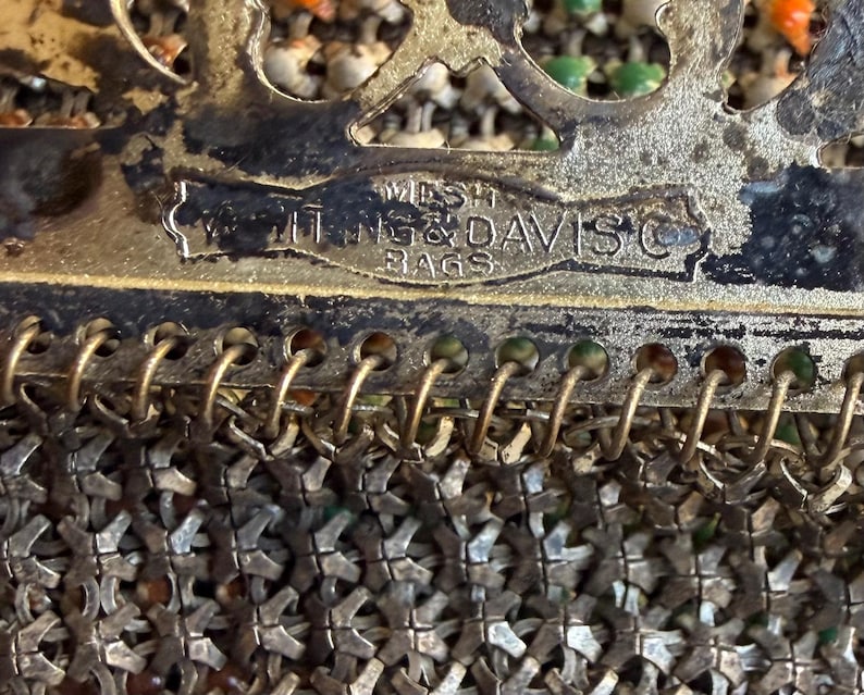 Op de afbeelding: Een vintage metalen mesh tas van Whiting & Davis Co. met een decoratief filigraan frame. Het metalen gaas bestaat uit kleine, in elkaar grijpende zilverkleurige schakels met subtiele oranje en groene emaille accenten zichtbaar in het weefsel.