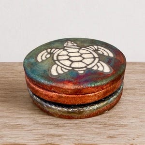 May include: A round ceramic lidded box featuring a white turtle graphic on the top. The lid and base display a blended glaze of metallic copper, deep teal, and dark red tones. The surface has a textured, mottled appearance with a glossy finish.