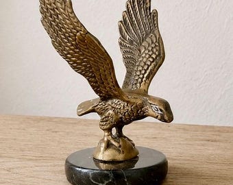 Op de afbeelding: Een koperen adelaarfiguur met wijd gespreide vleugels in een vliegende houding. De vogel staat op een kleine, ronde koperen basis die is bevestigd op een cirkelvormige, gepolijste zwarte marmeren voet met subtiele grijze aders erin.