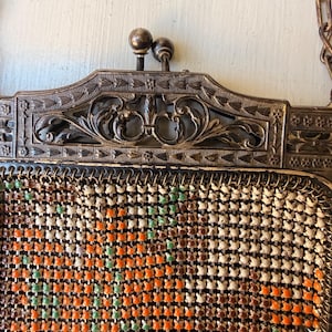 Op de afbeelding: Een handtas van metaalgaas in vintage-stijl met een rechthoekig frame, een sierlijk bloemmotief en een sluiting met twee balletjes. De tas is gemaakt van kleine, in elkaar grijpende metalen schakels in de kleuren oranje, bruin, cr&egrave;me en lichtgroen.