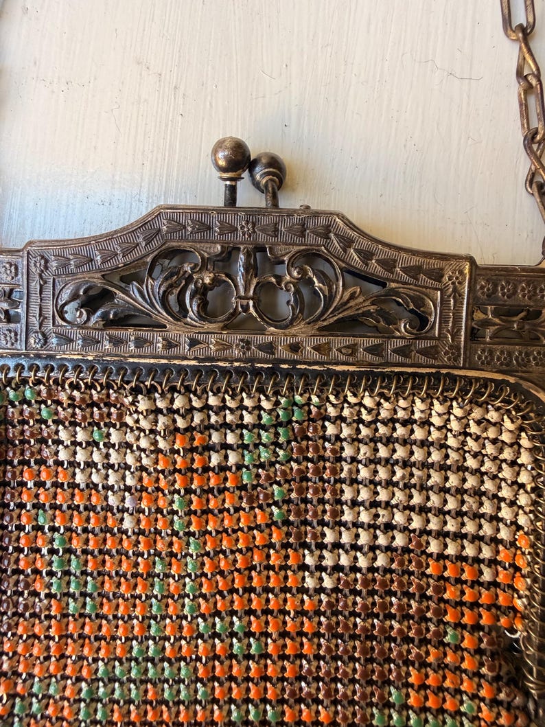 Op de afbeelding: Een handtas van metaalgaas in vintage-stijl met een rechthoekig frame, een sierlijk bloemmotief en een sluiting met twee balletjes. De tas is gemaakt van kleine, in elkaar grijpende metalen schakels in de kleuren oranje, bruin, cr&egrave;me en lichtgroen.