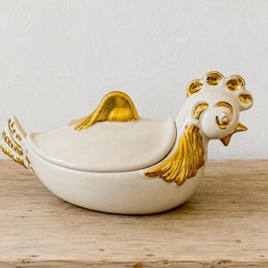 May include: A ceramic chicken-shaped lidded dish featuring a smooth off-white body with metallic gold accents on the comb, neck feathers, wing, and tail. The lid forms the back of the chicken, and the head is sculpted with a prominent gold crest.