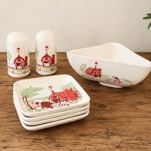 May include: A vintage ceramic dining set featuring a farm scene with red barns, cows, and green trees. The set includes two salt shakers, a serving bowl, and a stack of square plates. The pieces are white with a red, green, and brown design.