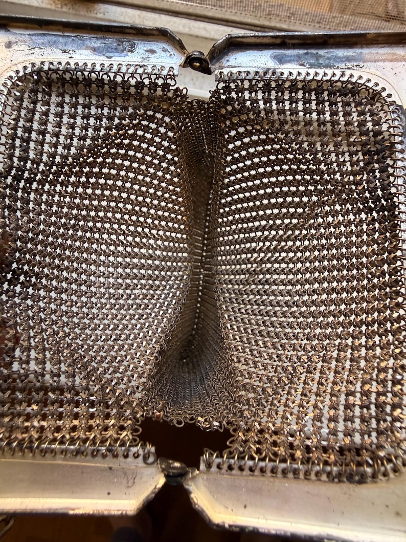 Op de afbeelding: Een open metalen mesh tas met een fijne, in elkaar grijpende zilverkleurige mali&euml;nkolderconstructie. Het interieur is verdeeld in twee afzonderlijke compartimenten door een centrale mesh scheidingswand, omlijst door een scharnierende metalen sluiting.