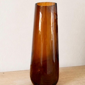 May include: A tall, tapered glass vase in a translucent amber brown color. The vase features a smooth, polished surface with a cylindrical shape that narrows slightly toward the top opening, showcasing a clear and reflective glass material.