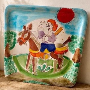 May include: Hand-painted ceramic tray with a whimsical design. The tray features a person riding a rocking horse under a red sun against a blue sky. The scene is framed by green trees and a green base. The tray has a square shape with rounded corners.