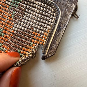 Op de afbeelding: Een vintage tasje van metaalgaas met een decoratief, sierlijk zilverkleurig frame met ingewikkelde reli&euml;fpatronen. De flexibele gaasbehuizing bestaat uit kleine, in elkaar grijpende schakels met een patroon van witte, oranje en lichtgroene kralen.