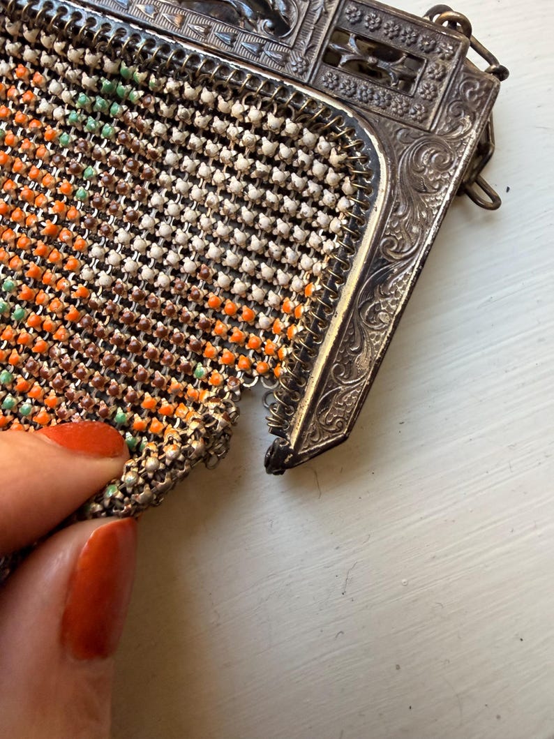 Op de afbeelding: Een vintage tasje van metaalgaas met een decoratief, sierlijk zilverkleurig frame met ingewikkelde reli&euml;fpatronen. De flexibele gaasbehuizing bestaat uit kleine, in elkaar grijpende schakels met een patroon van witte, oranje en lichtgroene kralen.