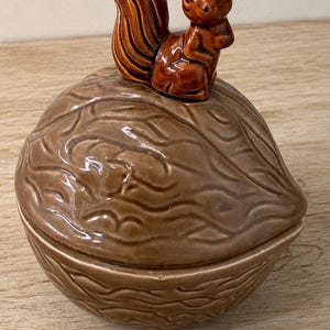 May include: A brown ceramic lidded container shaped like a walnut with a glossy finish. The lid features a sculpted brown squirrel figurine sitting upright on top, serving as a handle. The surface of the container has a textured, ridged pattern.