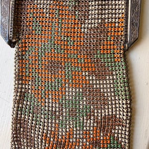 Op de afbeelding: Een vintage metalen mesh handtas met een bloemmotief van oranje, groene en cr&egrave;mekleurige emaille kralen. De tas heeft een sierlijk, gegraveerd zilverkleurig metalen frame met krulwerk en een geschulpte onderrand met hangende metalen schakels.