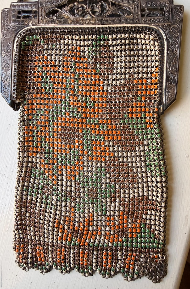 Op de afbeelding: Een vintage metalen mesh handtas met een bloemmotief van oranje, groene en cr&egrave;mekleurige emaille kralen. De tas heeft een sierlijk, gegraveerd zilverkleurig metalen frame met krulwerk en een geschulpte onderrand met hangende metalen schakels.