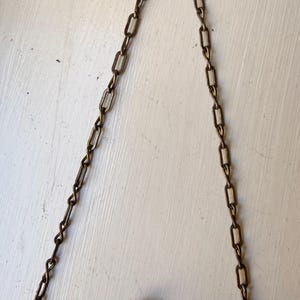 Op de afbeelding: Een metalen handtas met een gedetailleerd, donkerkleurig filigraan frame en een sluiting met twee knoppen. De tas hangt aan een lange ketting met rechthoekige schakels in een bijpassende antieke messing afwerking.