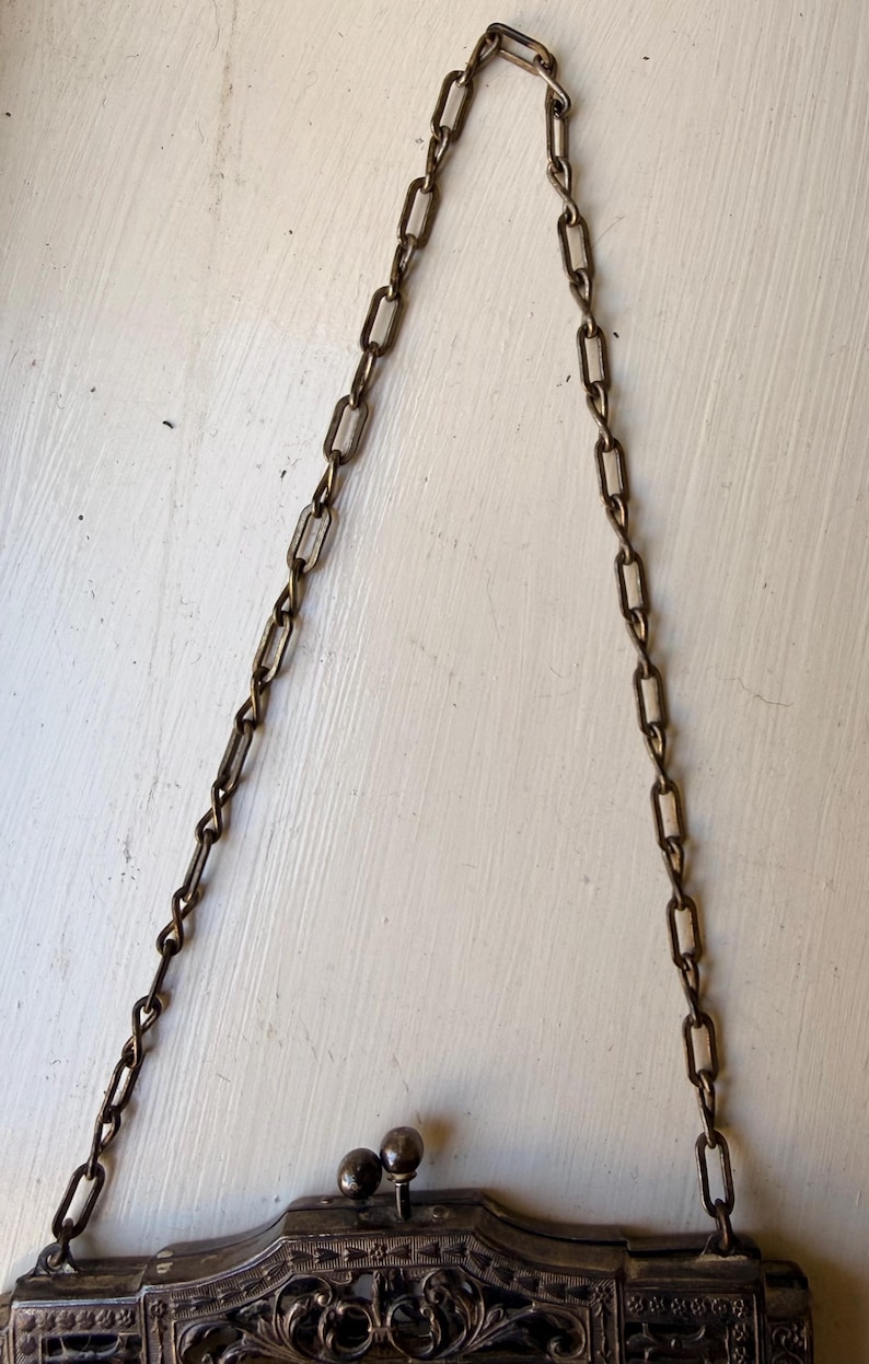 Op de afbeelding: Een metalen handtas met een gedetailleerd, donkerkleurig filigraan frame en een sluiting met twee knoppen. De tas hangt aan een lange ketting met rechthoekige schakels in een bijpassende antieke messing afwerking.