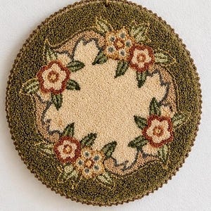 May include: A circular punch needle wall hanging featuring a textured olive green border and a cream-colored center. The design includes a wreath of embroidered flowers in shades of rust, peach, and green with dark green leaves.